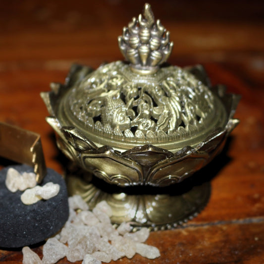 2 piece (top and bottom) ornate gold coloured incense holder, sitting on a wooden table, next to a pair of gold coloured charcoal tongs and a charcoal disc, with frankincense granules strewn around the base of the incense holder