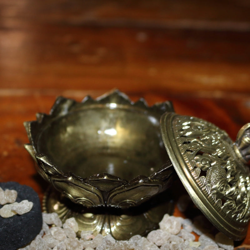 2 piece gold coloured ornate incense holder / censer on a wooden table next to a charcoal disc with frankincense granules strewn around the base of it