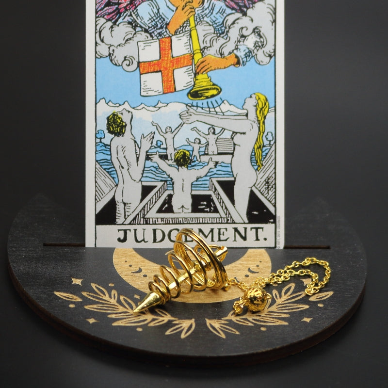 gold spiral pendulum sitting on a moon shaped piece of wood painted black with a crescent moon on it, with a groove cut down the middle of it, holding a judgement rider waite tarot card.