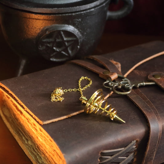 gold spiral pendulum sitting on a brown leather journal, in front of a black cast iron cauldron with a pentacle symbol on the front