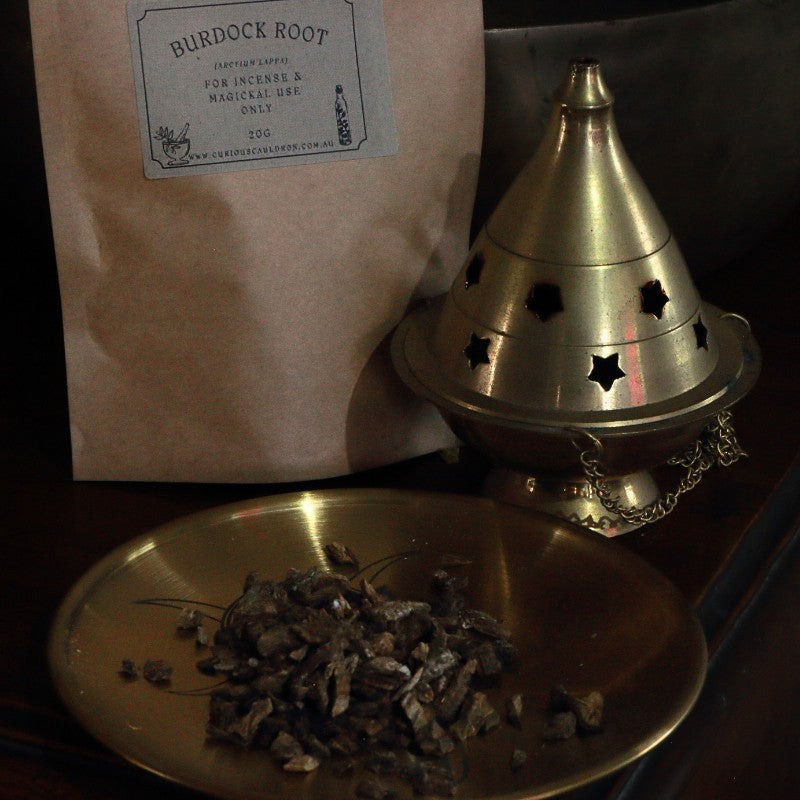 Brass incense burner with dried herbs on a metal plate, next to a bag labeled 'Burdock Root'.