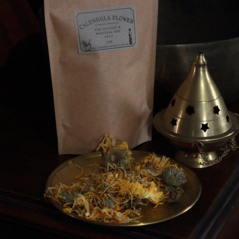 Dried calendula flowers on a gold plate with a bag labeled 'Calendula Flowers' and a brass incense burner.
