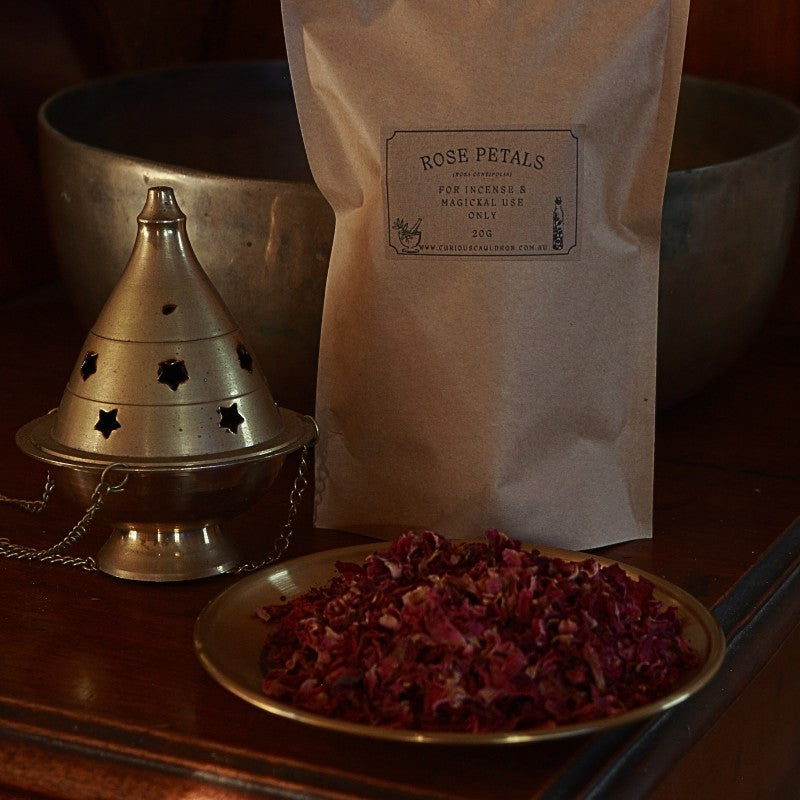 Bag of rose petals, bowl of dried rose petals, and metallic cone incense burner on a wooden surface.
