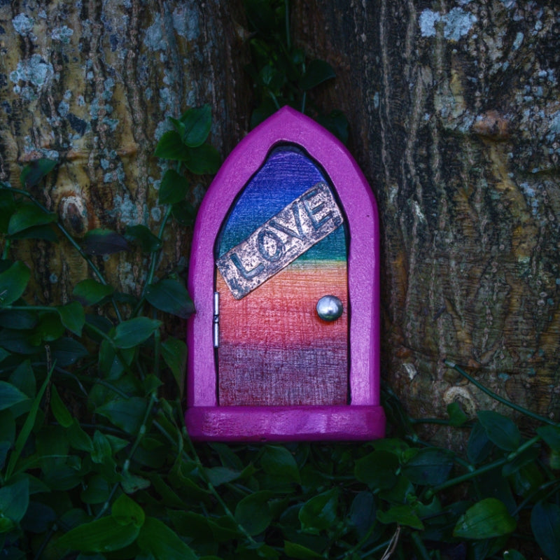fairy door made from wood, painted pink with a rainbow door and handmade metal hinges and door knob. The front of the door has an etched copper sign saying "love". the door sits between two trees atop dark green vines
