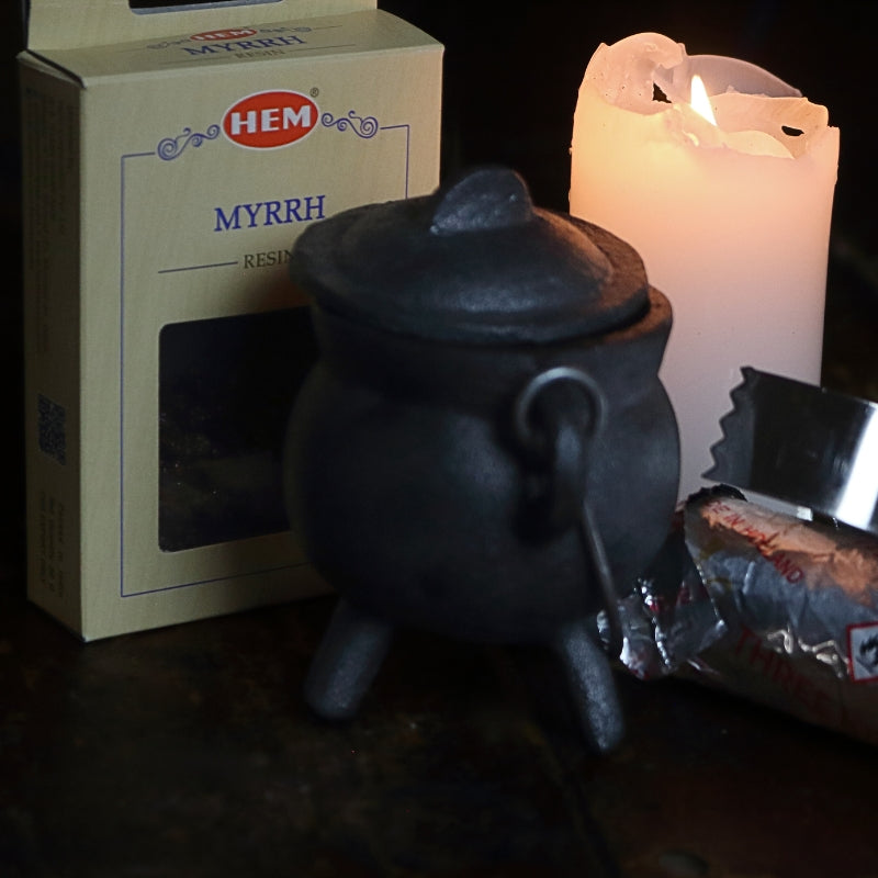 black cast iron cauldron in front of a beige coloured box of myrrh resin, next to a lit white church candle and a silver roll of charcoal discs and pair of stainless steel charcoal tongs on a wooden table