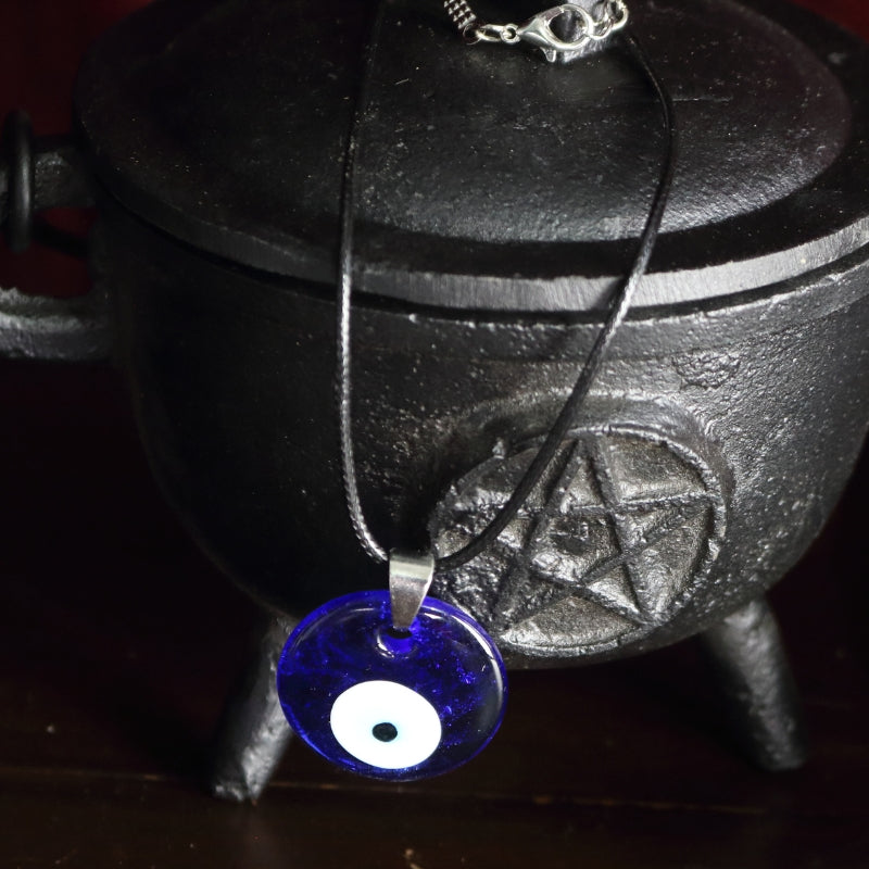 blue, white, light blue and black concentric circles  formed into an eye shaped bead, hung on a black cord with a silver clasp. The necklace hangs from a black cast iron cauldron with a pentacle on the front of it.