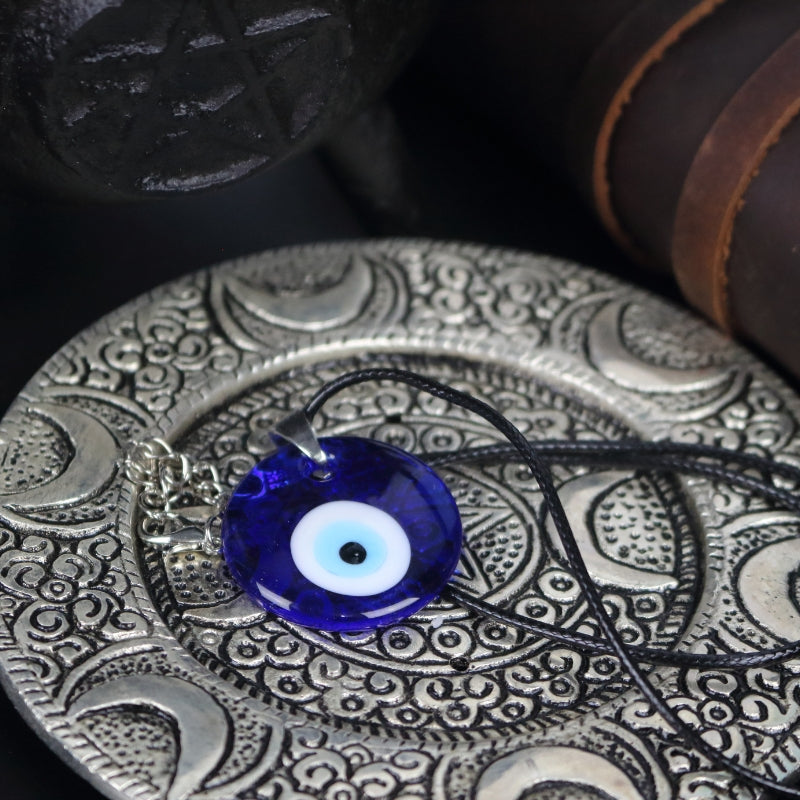 blue, white, light blue and black concentric circles  formed into an eye shaped bead, hung on a black cord with a silver clasp. The necklace sits on an ornate silver plate with moons embossed on it, in front of a black cast iron cauldron with a pentacle on the front of it.