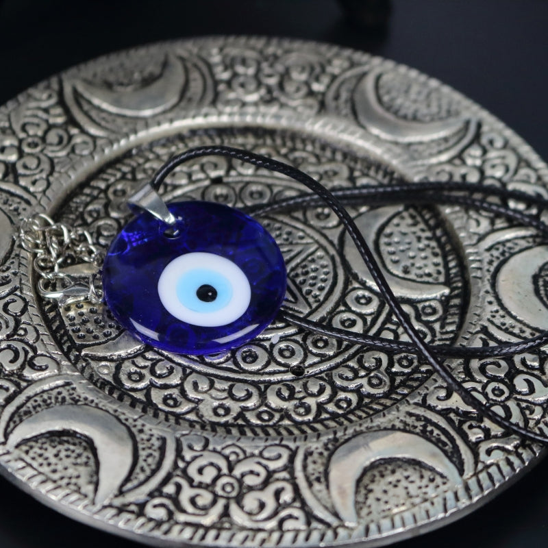 blue, white, light blue and black concentric circles  formed into an eye shaped bead, hung on a black cord with a silver clasp. The necklace sits on an ornate silver plate with moons embossed on it.