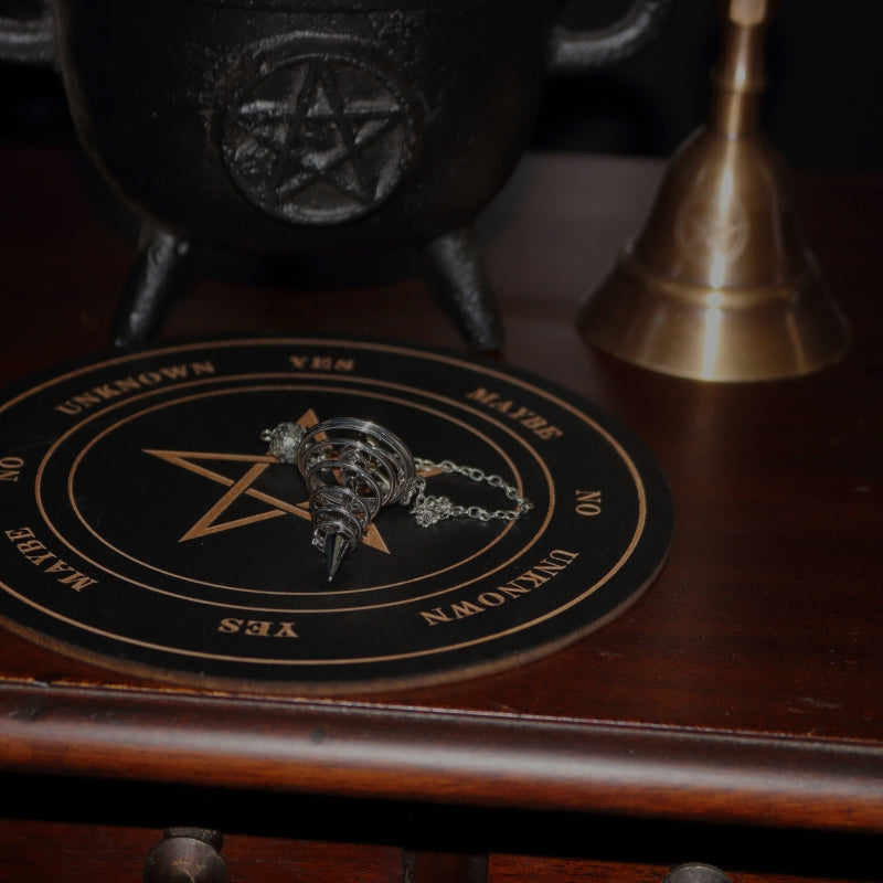 black round board for pendulum dowsing with the pentagram  in the centre  and yes, no, maybe and rephrase around the pentagram, sitting on am apothecary cabinet with a silver pendulum, brass altar bell and pentacle cauldron