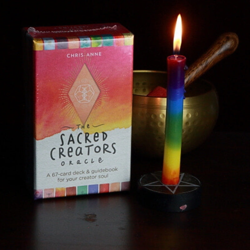 rainbow spell candle in a pentacle candle holder in front of a brass singing bowl, next to a pack of sacred creators oracle cards
