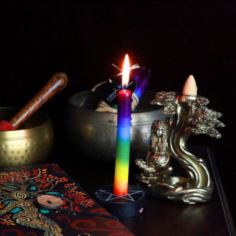 rainbow spell candle in a pentacle candle holder in front of a brass singing bowl, next to a mother earth goddess backflow cone burner and goddess journal, with a brass singing bowl in the background