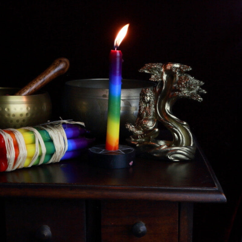 rainbow coloured candle in a spell candle holder on an apothecary cabinet next to brass singing bowls and a goddess mother earth backflow incense burner