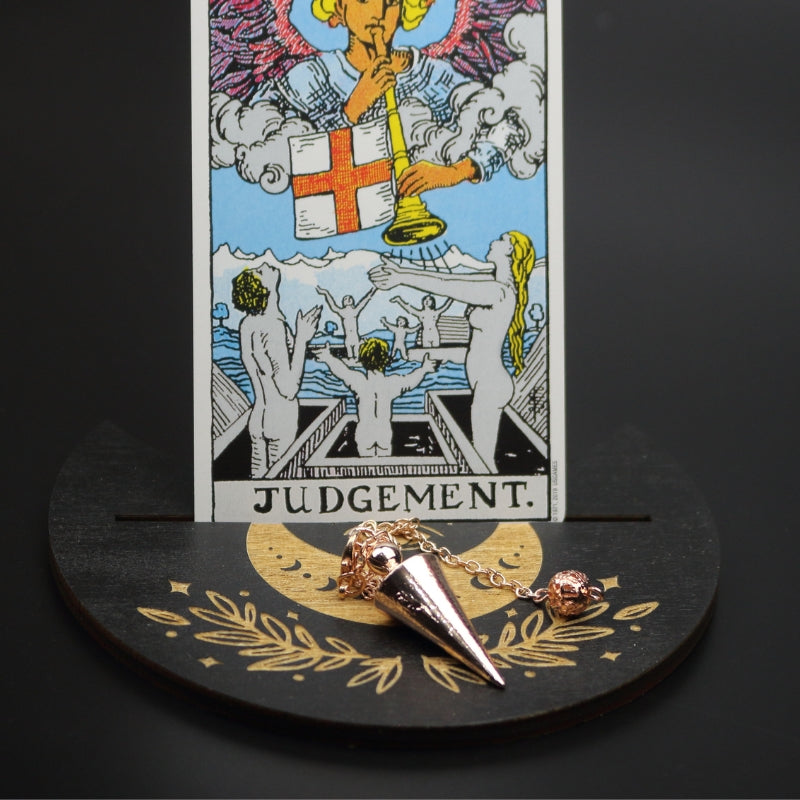 rose gold conical pendulum sitting on a moon shaped piece of wood painted black with a crescent moon on it, with a groove cut down the middle of it, holding a judgement rider waite tarot card.