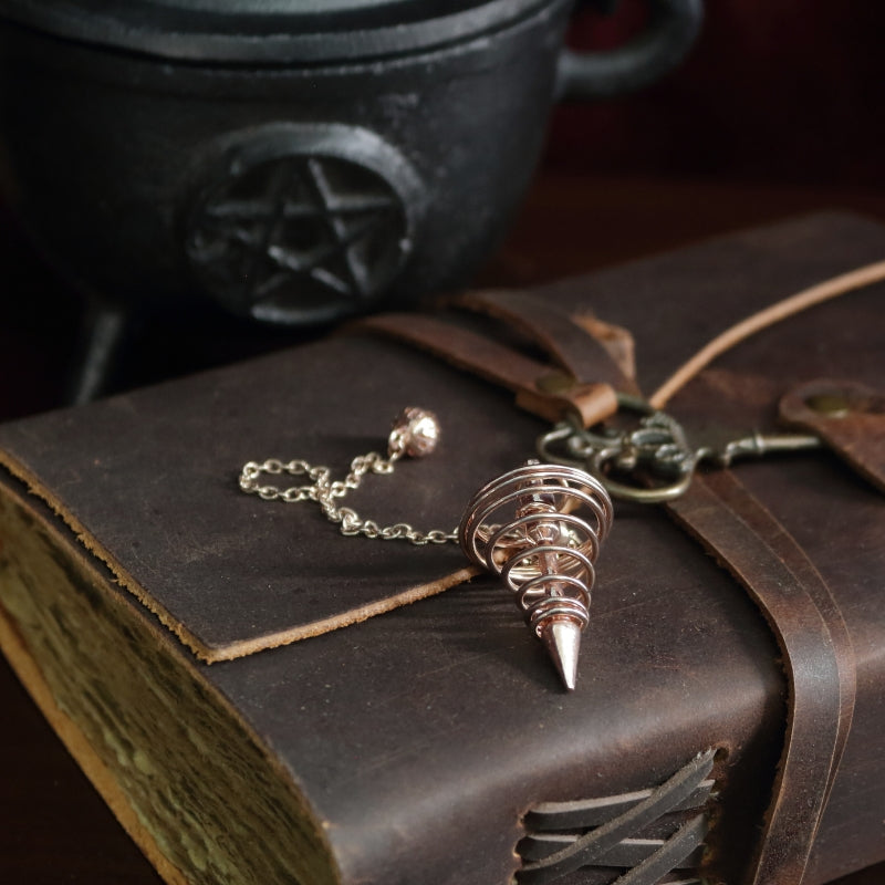 rose gold spiral pendulum sitting on a brown leather journal, in front of a black cast iron cauldron with a pentacle symbol on the front