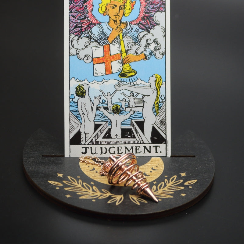 rose gold spiral pendulum sitting on a moon shaped piece of wood painted black with a crescent moon on it, with a groove cut down the middle of it, holding a judgement rider waite tarot card.