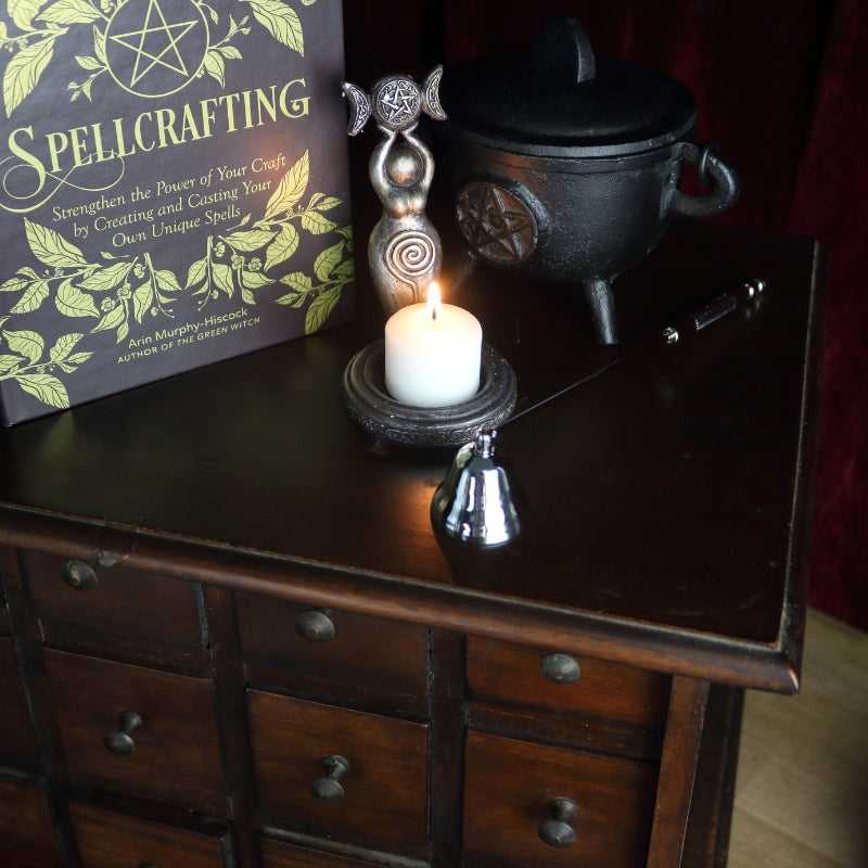 apothecary cabinet with a spellcrafting book on top, a black cast iron cauldron with a pentcle design on front, a silver candle snuffer, candle holder in the shape of a goddess holding a triple moon.
