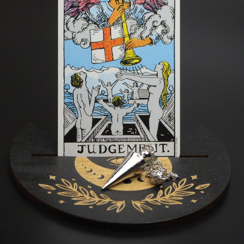 silver conical pendulum sitting on a moon shaped piece of wood painted black with a crescent moon on it, with a groove cut down the middle of it, holding a judgement rider waite tarot card.