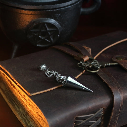 silver conical pendulum sitting on a brown leather journal, in front of a black cast iron cauldron with a pentacle symbol on the front