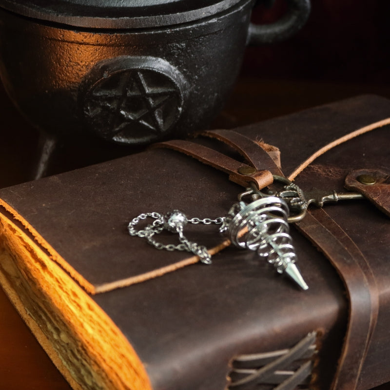 silver spiral pendulum sitting on a brown leather journal, in front of a black cast iron cauldron with a pentacle symbol on the front