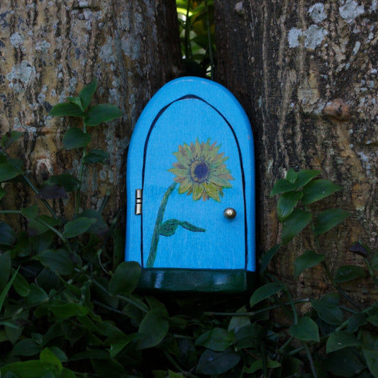 blue flower fairy door with a painted yellow sunflower and green doorstep- silver hinge and doorknob, sitting in front of tree