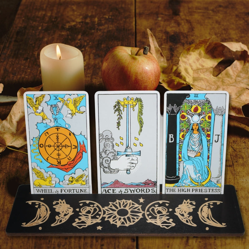 3 tarot cards in a black tarot card holder on a wooden table with autumn leaves, an apple and a lit white pillar candle in the background