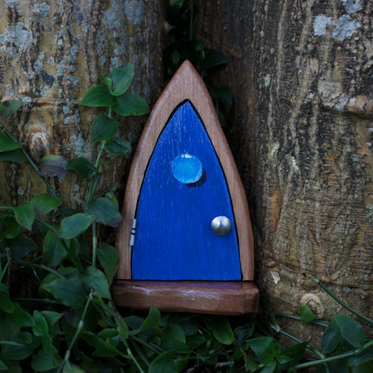 Handmade wooden fairy door with a water drop painted on the front, standing in front of a tree