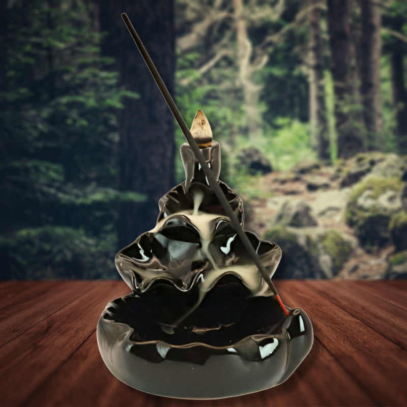 Ceramic Waterfall Backflow Incense Cone Burner with a sitck incense in front and smoke billowing down the front in front of a rainforest background