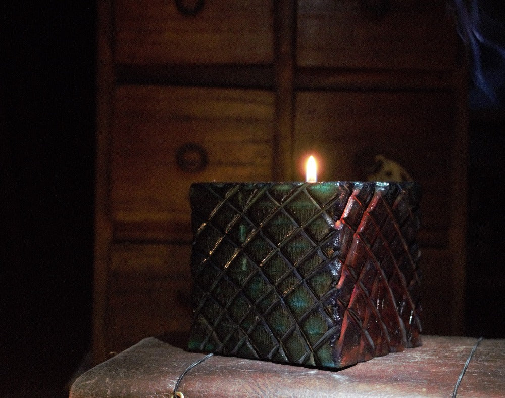 blue and red sides of multicoloured wooden square candle holder with diagonal lines etched to resemble dragon scales. Containing a white tea light candle, on a leather journal, in front of an apothecary chest