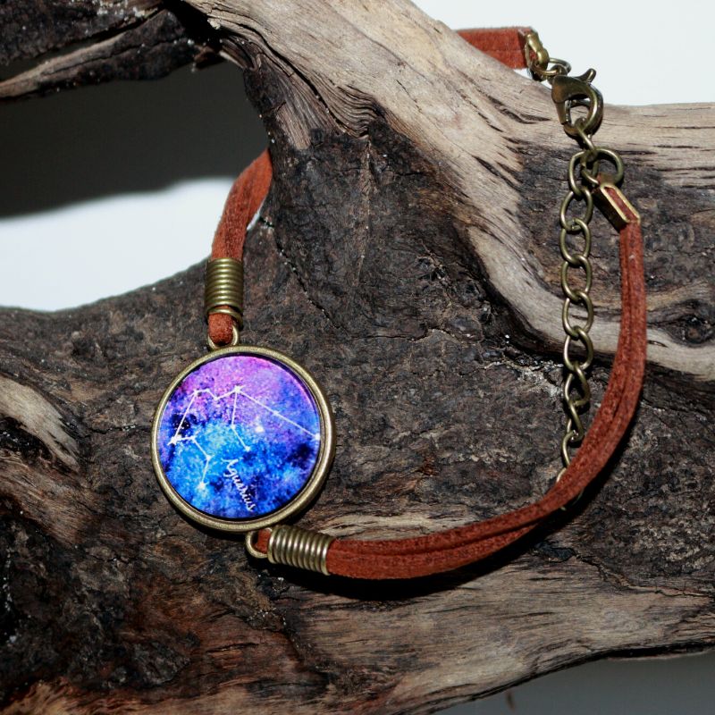 leather adjustable bracelet with a glass cabochon in the centre featuring an image of the aquarius constellatin line drawing on a blue and purple background, hanging on a piece of drift wood