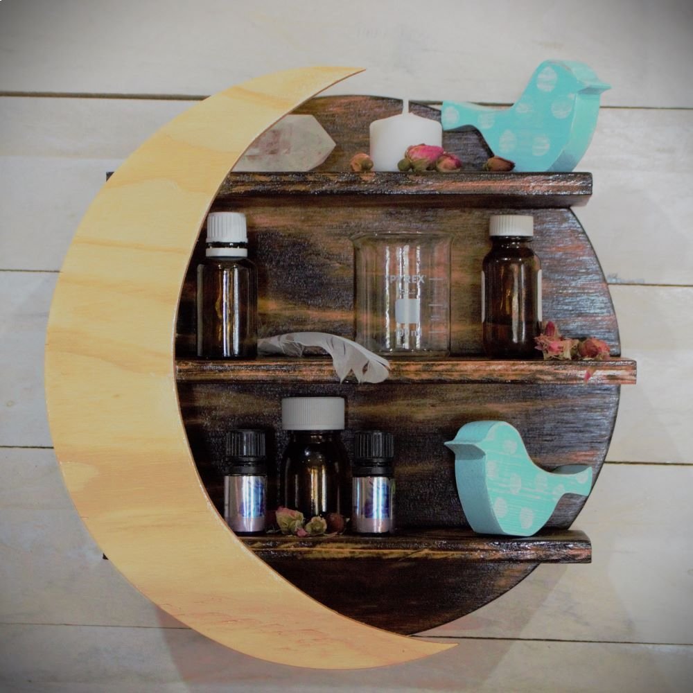 wooden crystal moon shelf with light wooden crescent moon in front of 3 shelves, holding crystals, glass beakers, candle, blue and white spotted ornamental birds and essential oil bottles, on a white slat wall