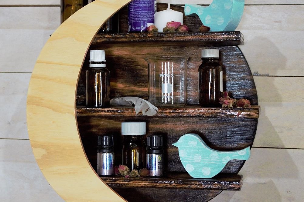 wooden crystal moon shelf with light wooden crescent moon in front of 3 shelves, holding crystals, glass beakers, candle, blue and white spotted ornamental birds and essential oil bottles, on a white slat wall