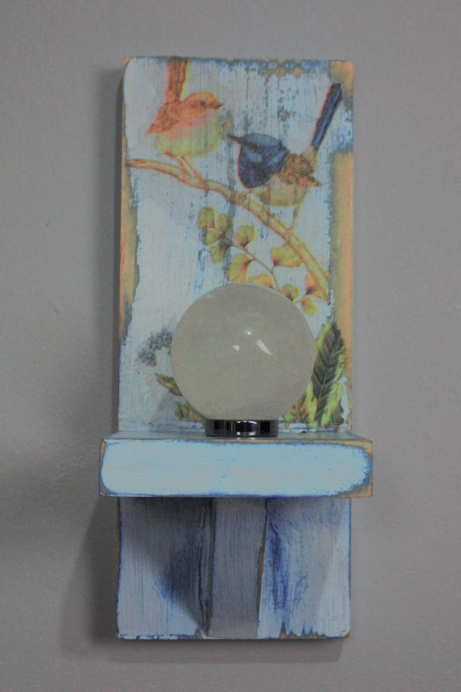 clear quartz crystal ball atop a hematite ring sitting  on a distressed blue and white wooden wall mounted candle or ornament shelf decorated with vintage style brown and blue wrens sitting on a branch with green ferns and blue flowers