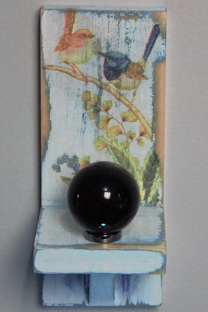 Black obsidian crystal ball sitting on a hematite ring atop a distressed blue and white wooden wall mounted candle or ornament shelf decorated with vintage style brown and blue wrens sitting on a branch with green ferns and blue flowers
