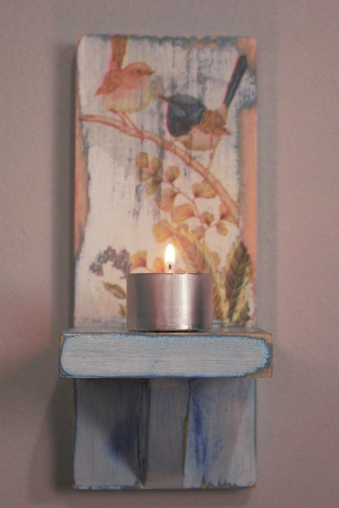 metal tea light  candle sitting on a distressed blue and white wooden wall mounted candle or ornament shelf decorated with vintage style brown and blue wrens sitting on a branch with green ferns and blue flowers