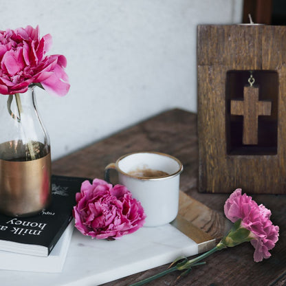 cross wooden tealight candle holder  solid hardwood with white tea light in top, next to a board with books, coffee and pink carnations, on wooden table with white background wall