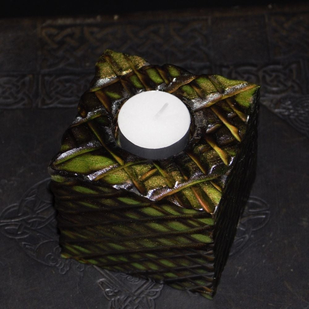 green and gold wooden square candle holder with diagonal lines etched to resemble dragon scales. Containing a white tea light candle, on a black embossed background