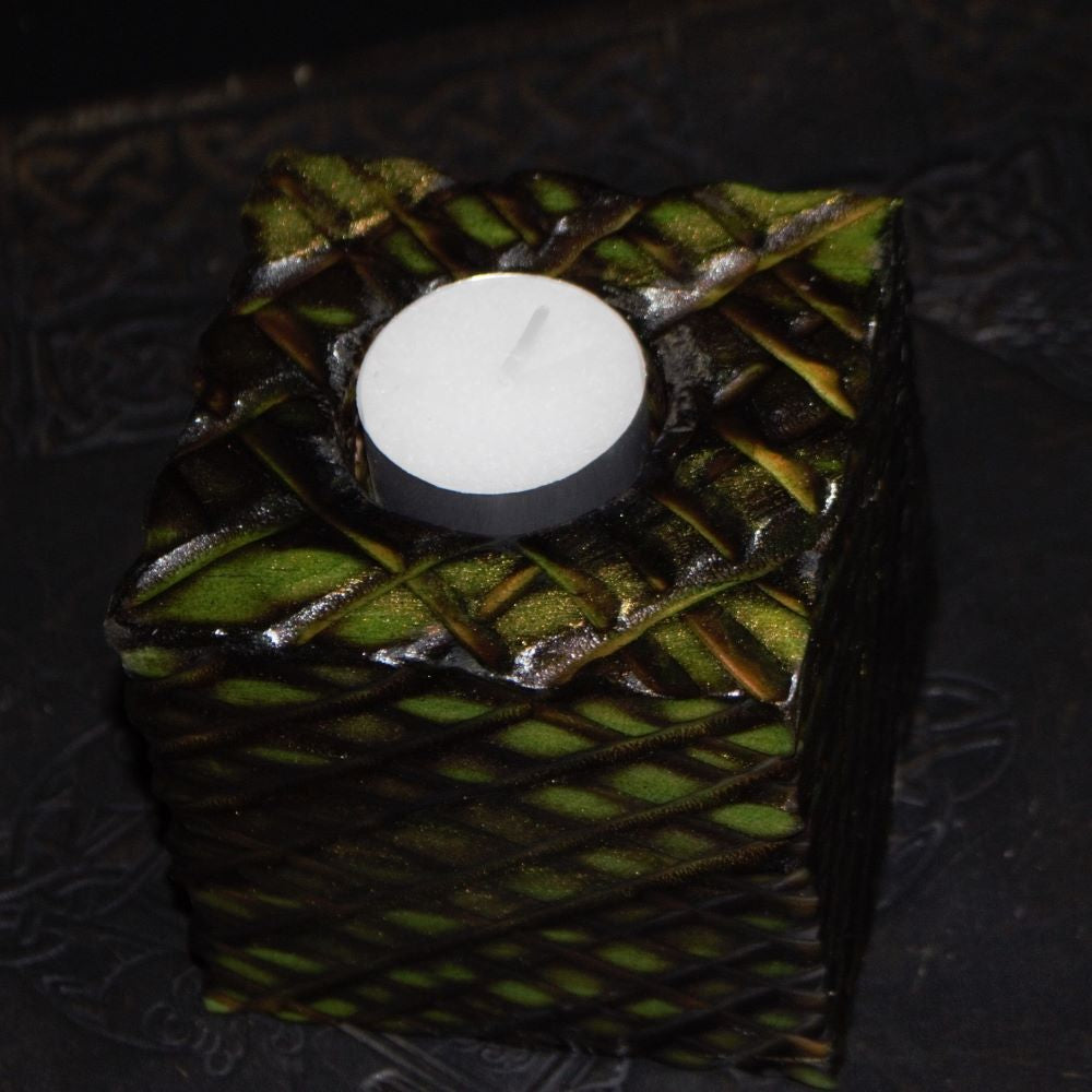 green and gold wooden square candle holder with diagonal lines etched to resemble dragon scales. Containing a white tea light candle, on a black embossed background