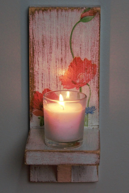 glass white votive candle sitting on a red and white wooden wall mounted candle or ornament shelf decorated with red and blue flowers