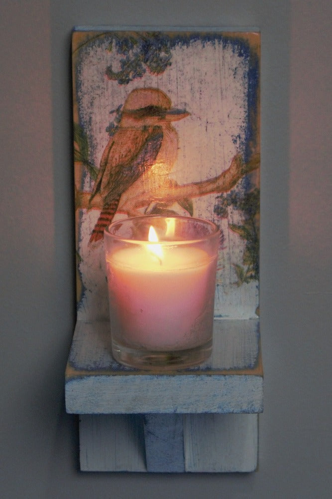 glass white votive candle on a distressed blue and white wooden wall mounted candle or ornament shelf decorated with vintage style kookaburra sitting on a branch with red and blue flowers