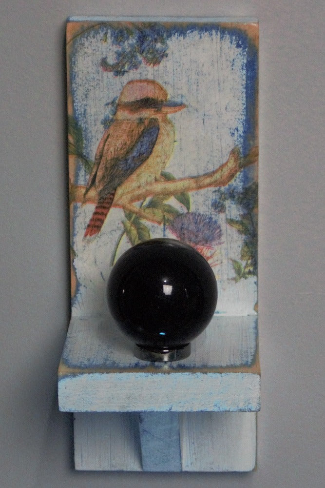 Black obsidian crystal ball sitting on a hematite ring atop a distressed blue and white wooden wall mounted candle or ornament shelf decorated with vintage style kookaburra sitting on a branch with red and blue flowers