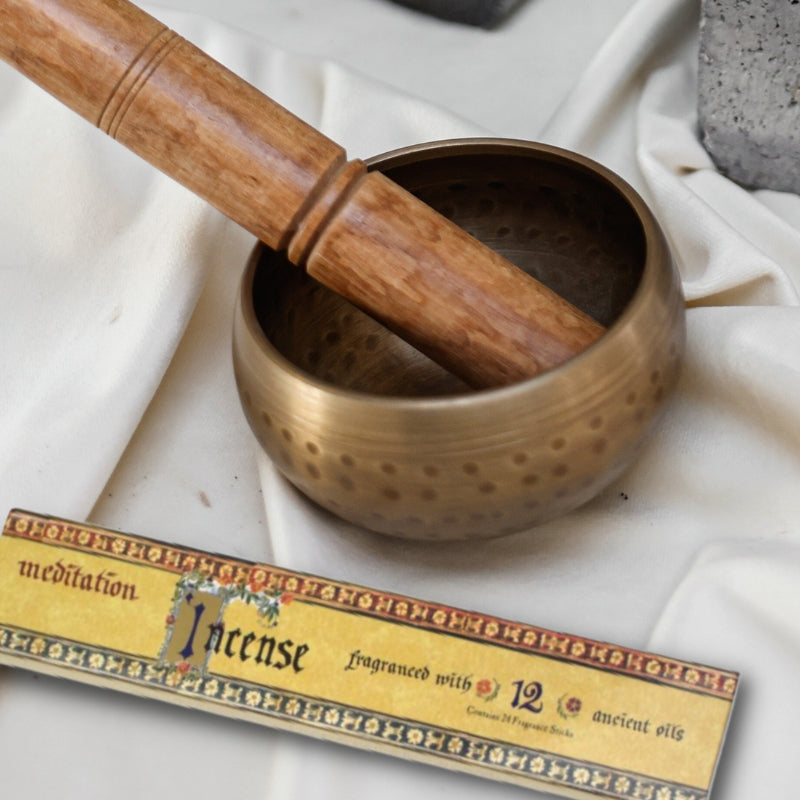 beige coloured linen cloth with a brass singing bowl on top, with a wooden striker in bowl. A packet of meditation incense sits in front of the singing bowl