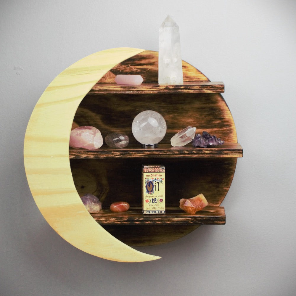 wooden crystal moon shelf with light wooden crescent moon in front of 3 shelves, holding crystals and meditation fragrance essential oil, on a grey wall