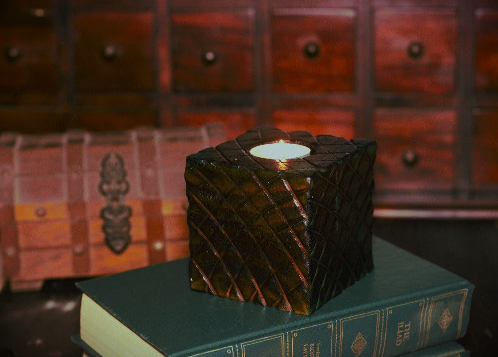 Red wooden square candle holder with diagonal lines etched to resemble dragon scales. Containing a white tea light candle, on a stack of books , in front of an apothecary chest and wooden treasure chest
