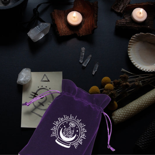 purple tarot bag with white moon and crystal printon table with a rolled beeswax candle, clear quartz, a seashell and candles