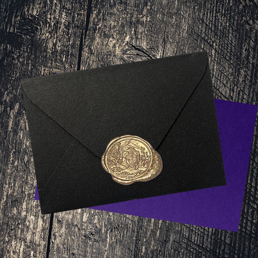 gold wax seal of a raven on a black envelope , sitting on top of a purple envelope, on a wooden table