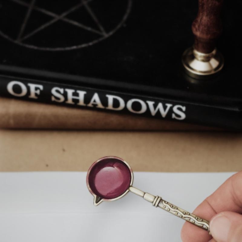 wax melting spoon with pink wax being held over a white envelope, in front of a book of shadows and leather journal